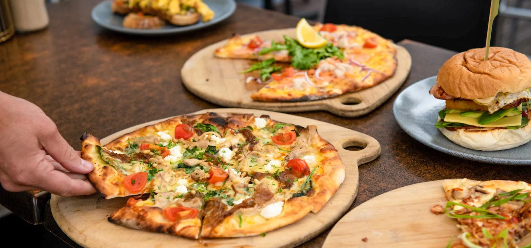 Close up view of pizzas and burgers on a table. A person is taking a slice